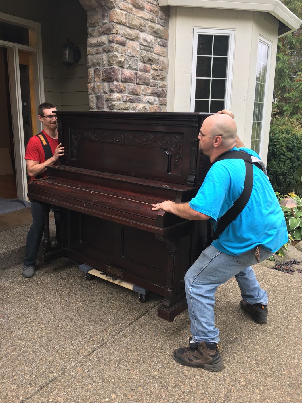 Two Confidence movers walking an upright piano out of a front door